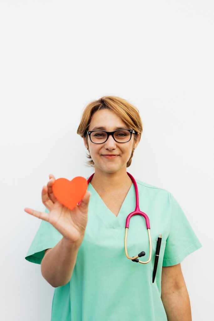 pexels-photo-5207117 Smiling healthcare professional holding a heart symbol, standing confidently with a white background.