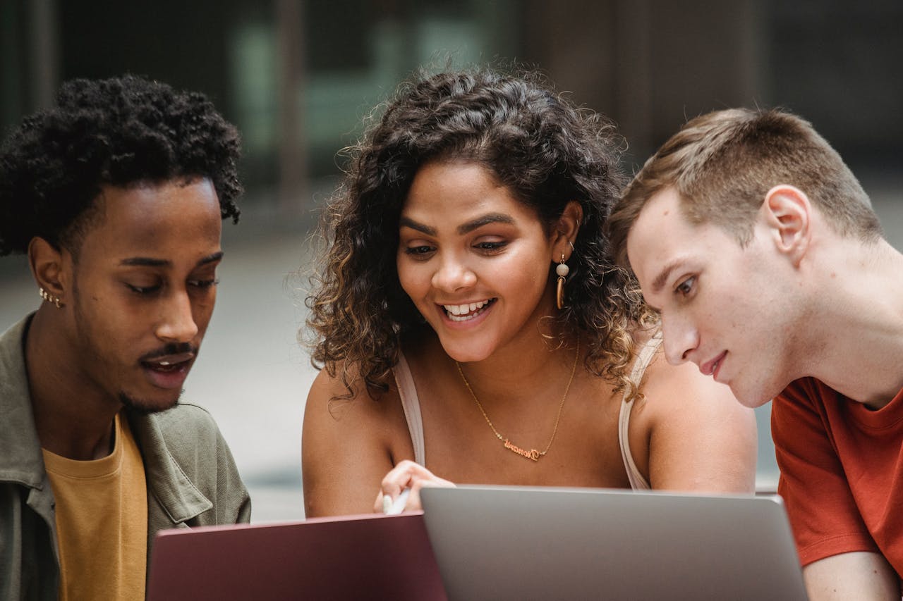 Three young adults of different ethnicities happily collaborating on laptops outdoors, promoting teamwork and diversity.