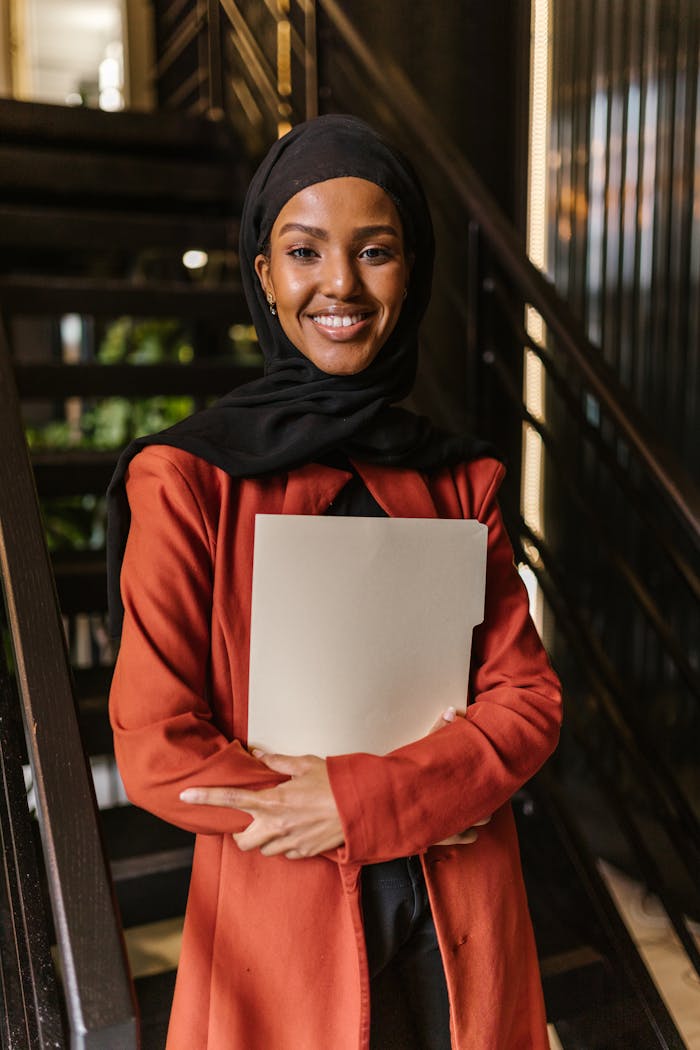 Professional woman in hijab holding folder, smiling in modern office setting.